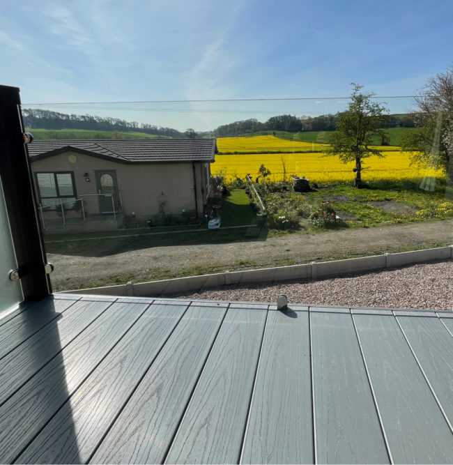 Edge of a balcony with a glass handrail, looking out into the countryside with yellow sunflowers trees and blue skys